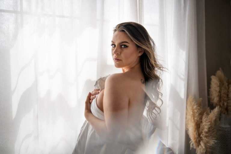 woman holding white sheet standing by window with hair blowing and sunlight shining through the sheer curtain